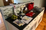 Author’s photo of a kitchen counter with piles of papers, a laptop, red purse, basket of apples