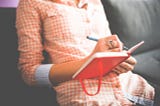 A photo of a woman without the head visible, dressed in bright red checkered dress. She is writing in a hot pink notebook.