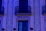 A side of a building with windows with balconies. There is decorative ornamentation on the walls, and the entire building is lit up by a blue light. One of the balconies has a string of lights on it.
