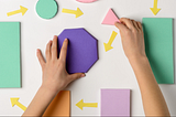 Two hands are seen arranging colorful geometric shapes on a white surface. The shapes include a purple octagon, a pink triangle, and several rectangles in green, orange, and purple. Yellow arrows, also made from paper, point in various directions, indicating a process or sequence. The arrangement suggests an exercise in design, planning, or problem-solving.Female hands precisely arranging a project plan with sticky notes and arrows.