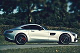 A sexy Silver Mercedes in profile against a backdrop of a hedge and green trees.