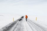 Wide view of a snowfield and a partially plowed road in the middle. A person is walking away from the viewer on the snowy road.