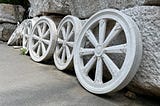 Wheel of Buddhist Dharma, a symbol of teaching in Buddhism. White marble wheels are set in a row, leaning against large boulders, with a statue of a white stork in the background. Installation at a Buddhist temple. Image credit: Oxana via Adobe Stock.