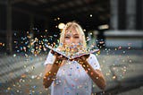 A blonde woman holding a book open in front of her face, blows rainbow confetti off the pages toward the viewer.