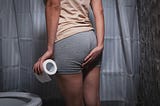 A person in grey shorts holding their butt crack with one hand and a roll of toilet paper in the other
