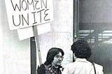 Lynn Fraser being interviewed by a reporter during a demonstration holding a sign Uppity Women Unite