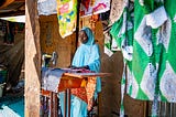 A woman works outside her home at a sewing machine table surrounded by a variety of colorful material hanging overhead and behind her.