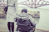 Anna and her brother pictured overlooking a bridge in South Queensferry. They are both wearing grey coats, and her brother sits in an electric wheelchair