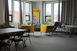 A view of the presentation room at Code & Supply Workspace. Some folding tables and chairs are to the left while the Uptime Lounge sits in the far corner. A bicycle lays against the window next to a plant.