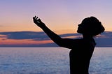 Silhouette of a young female against a sunset. She has a hand raised to the sky.