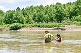 A man in waders stands waist deep in the Cuyahoga River in a wooded location. The treeline in the distance is bright green. Beside him is a shorter colleague and the waterline is up to her torso.
