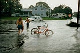 young people play in flooded street, with a car in background immersed in floodwaters. Bicycle in foreground.