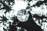 A hand holds a glass ball in a forest underneath some trees