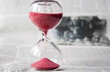 Clear glass hourglass with red sand running through it against a greyed out backdrop of printed paper.