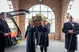 A wicker coffin garlanded with flowers being carried into a funeral service by four pallbearers. Only the two at the front are visible: a middle-aged woman and a younger women who might be her daughter.