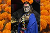 A “Catrina” wearing colorful death/skeleton makeup — she has marigolds in her dark hair and is holding a black umbrella. She’s wearing a black top, a blue cape and a colorful skirt with beaded necklaces and earrings.