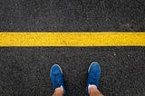 person looking down at shoes standing before a yellow line