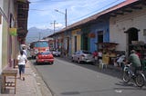 A street in Granada, Nicaragua showing low-rise multi-coloured buildings, a colourful bus, a taxi, people moving around and a volcano in the distance.