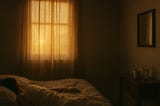 Soft amber light filters through curtains onto a bed where a woman lies still beside a small table.