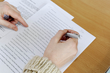 Hands in a sweater hover over a typed piece of paper with edit marks on it all over a light wooden surface. The left hand points at a line while the right hand holds a pen.