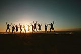 Ten people in a line, simultaneously jumping into the air joyfully, on a beach, at sunset.