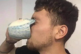 A person drinking from a blue mug with an unusual design, where the rim of the cup is curved inward, making it awkward to drink from. The mug’s shape blocks the person’s face, giving a humorous and impractical impression.