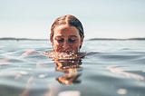 Woman emerges from the water, eyelids lowered and sun shining down.