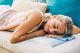 A woman with blond hair lays on the couch with her head and hands resting on an open notebook.