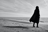 A hooded figure stands on a sand facing the water. The figure is alone and casts a long shadow.