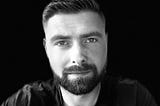 A black and white close-up portrait of a serious-looking man with short dark hair, a full beard, and intense eyes, gazing directly at the camera against a dark background.