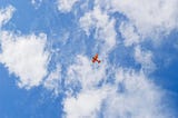 Small red plane against wide clouds in blue sky photographed from below