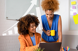 A woman sitting down looks at a computer and talks to another woman who is standing up.