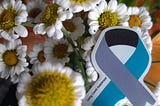 Blue and grey Long Covid awareness ribbon pin badge next to daisies