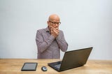 Image shows bald man in glasses standing behind long, wooden desk with laptop, computer mouse, and tablet on it. He stares horrified at the laptop screen with hands clasped over mouth.