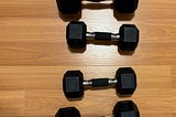 Four hexagonal dumbbells arranged in two columns on a wooden floor. The weights are labeled as 15 pounds and 10 pounds, with the 15’s  weight wrapped in tape on the handle for added grip. The dumbbells feature rubber-coated ends and metal handles with knurled grips.