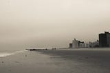 Moody photo of the ocean on the left & apartment buildings on the right, separated by a strip of sandy beach at Coney Island.