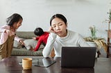 Young mom at a computer, while kids run rampant in the background. Mom’s eyes are closed, and she’s propping her head up with her arm leaning on the desk. She’s tired!