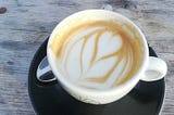 A half-drunk fancy coffee in a white china cup on a black dish, sitting on a wooden table