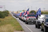 A convoy of pickup trucks flying “Trump 2020” flags stretches down a Texas road.