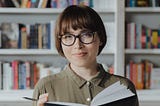 A woman with glasses looking up from a book with a pensive expression