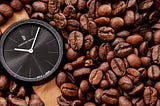 A watch with a light-brown vegan leather strap laying in a pile of coffee beans. The time on the watch shows approximately 10:07, and the watch case is made from recycled coffee grounds.