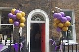 An open door in an arched doorway with a bundle of balloons floating on either side attached to the railings and bunting decorating the railings