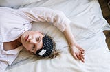 Woman spread out on bed with sleeping eye mask on her forehead looking done.