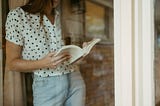 Linea reading a book through a window. She is wearing a white and black polka dot shirt.