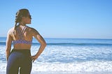 A woman stands on the beach in athletic wear staring out into the ocean with a renewed sense of confidence