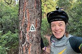 The author, Patty George, smiling in her Team PHenomenal Hope uniform and bicycle helmet, standing next to a tree with the Colorado trail logo. She is wearing a cycling cap with the words “Let Me Be Your Lungs”