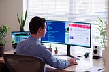 A professionally dressed man works at home with double computer screen and laptop on his desk with plants facing window