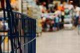 A close up picture of a blue shopping cart making it’s way to the check out aisle. The check out aisle in the background is blurred.