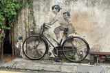 Penang street art — two kids on a bicycle — painted on a concrete wall with a real bicycle leaning against the wall.