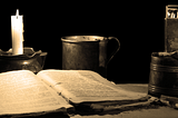 Tampo de uma mesa empoeirada com uma vela à esquerda, um livro antigo aberto e uma caneca de metal